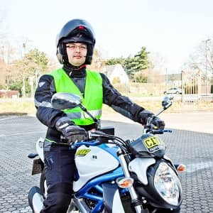Ein Mann sitzt auf einem Motorrad auf einem Parkplatz. Er trägt eine volle Motorradmontur und dazu eine Warnweste und einen Helm.