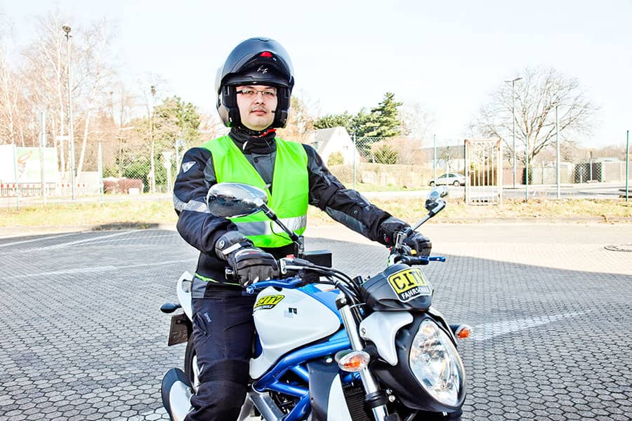 Ein Mann sitzt auf einem Motorrad auf einem Parkplatz. Er trägt eine volle Motorradmontur und dazu eine Warnweste und einen Helm.