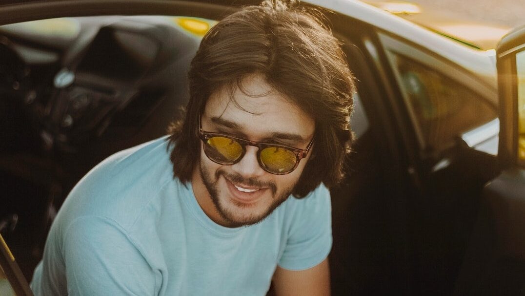 Ein junger Mann mit Sonnenbrille und schulterlangem, dunklem Haar sitzt in einem Auto und lehnt sich lächelnd aus der geöffneten Tür. Er trägt ein helles T-Shirt und blickt entspannt zur Seite. Die Szene ist von warmem Sonnenlicht beleuchtet.