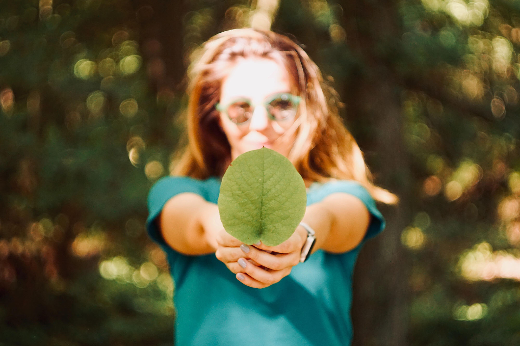 Eine Frau mit Sonnenbrille steht vor einem grünen Laubhintergrund in der Natur und hält ein rundes, grünes Blatt in die Kamera.