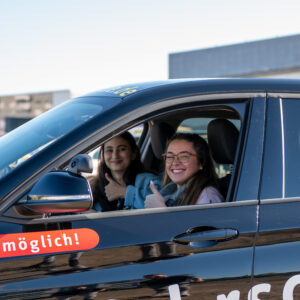 Zwei junge Frauen sitzen in einem Auto und zeigen mit einem breiten Lächeln einen Daumen hoch in die Kamera.