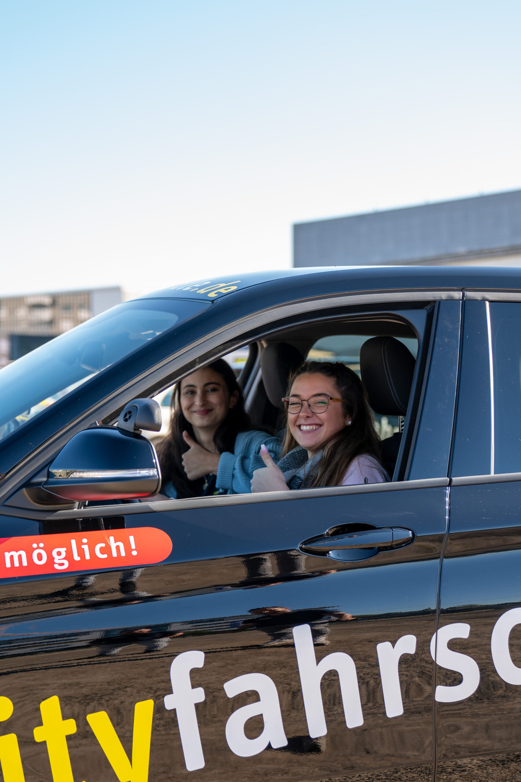 Zwei junge Frauen sitzen in einem Auto und zeigen mit einem breiten Lächeln einen Daumen hoch in die Kamera.