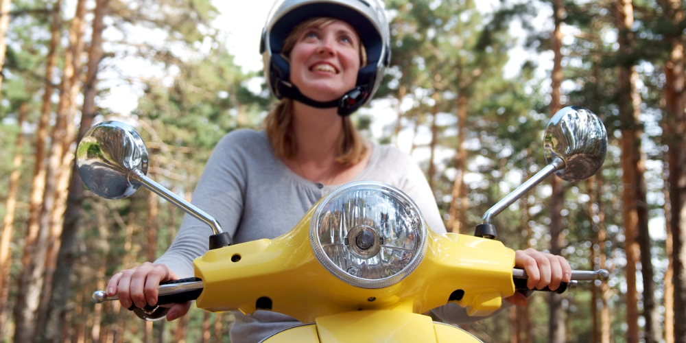 Frau sitzt auf Vespa, fährt durch den Wald und lächelt.